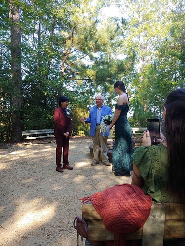 A Chinese man in a dark burgundy suit stands with a white woman in a long dark green dress. She is holding a large white bouquet. A wedding officiant in a blue suit jacket stands between them.