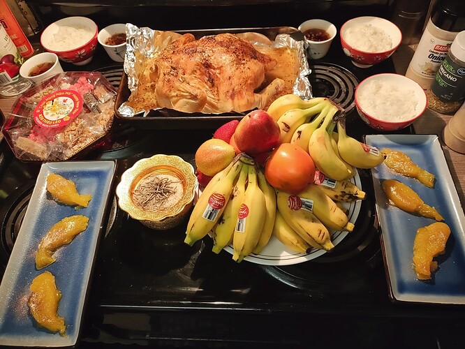 Stovetop with an incense bowl filled with uncooked rice and spent incense, 3 bowls of cooked rice, 3 cups of tea, a roast chicken, a tray of candies, 2 long blue trays with 3 mango agar jellies shaped like fish with glitter on them, and a plate stacked with mini bananas, persimmons, strawberries, mandarin oranges, and forelle pears.