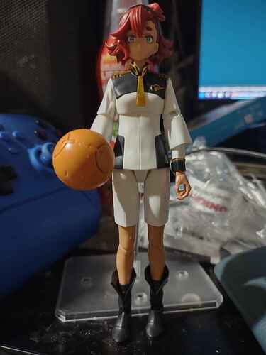 a model kit of the main character of Mobile Suit Gundam: The Witch From Mercury. Her name is Suletta Mercury. She is a middle eastern bisexual woman. She's wearing her special uniform, primarily white and black with gold trimmings. She has red hair and light brown skin. She is wearing zip up boots that are not fully zipped up. She is also holding an orange Haro, a spherical robot that appears in many Gundam works and universes.