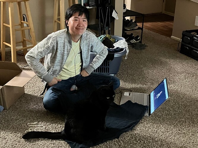 A Chinese man sits next to a laptop with his hands on his hips and a strained closed-mouth smile. A black cat is sitting on the t-shirt he was folding.